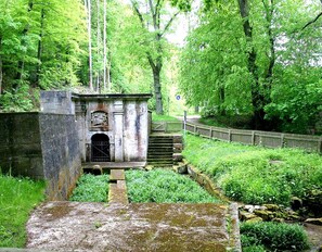 A stone structure with a metal gate, surrounded by lush greenery and a wooden fence. - Welcome to the "FUCHSREVIER"  (Erkerode)
