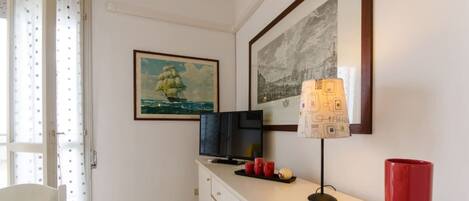 A room with a white dresser, a television on top, a lamp with a patterned lampshade, and two framed pictures on the wall.