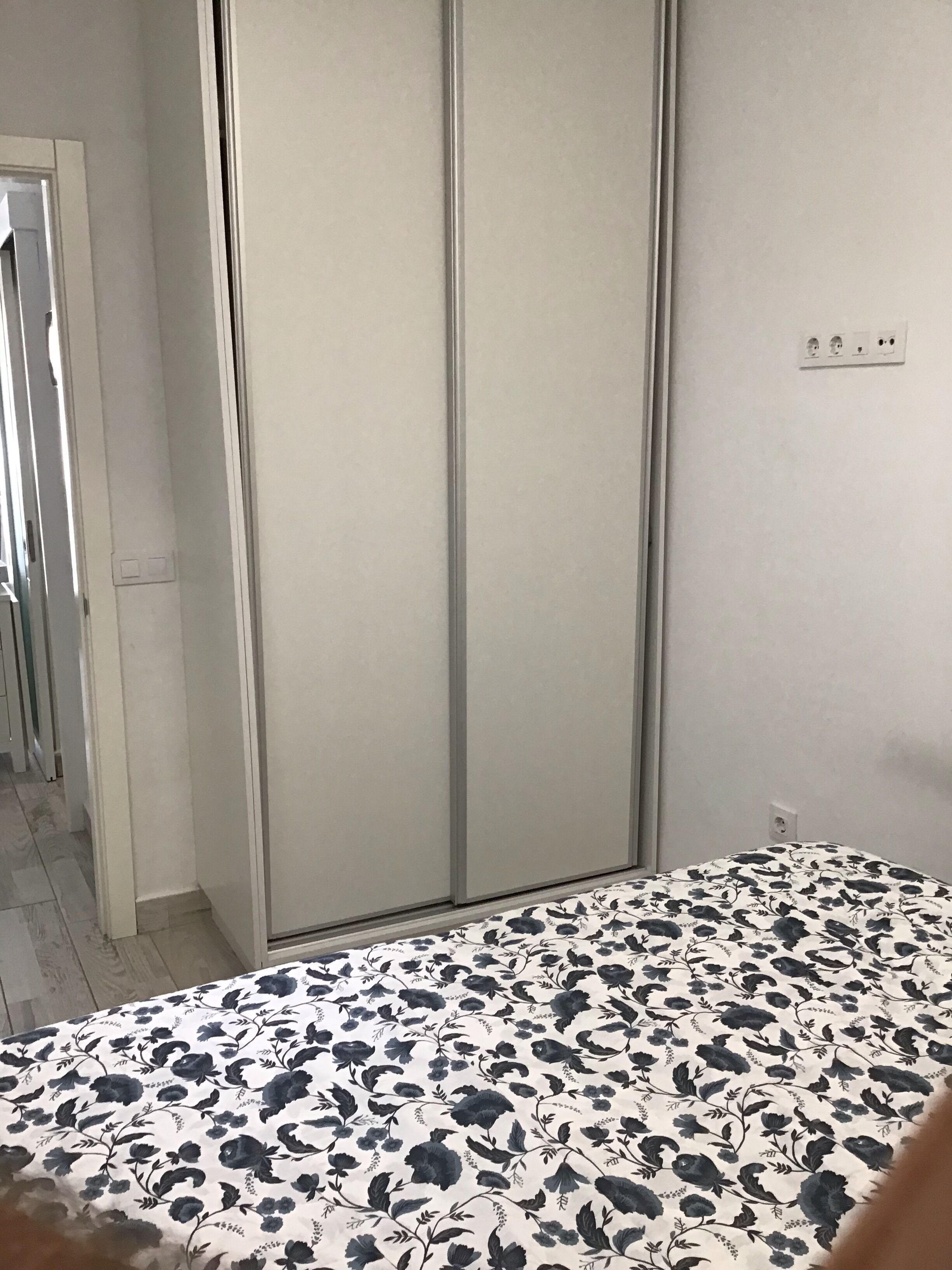 A bedroom with a large wardrobe, a patterned bedspread, and a visible electrical outlet on the wall.