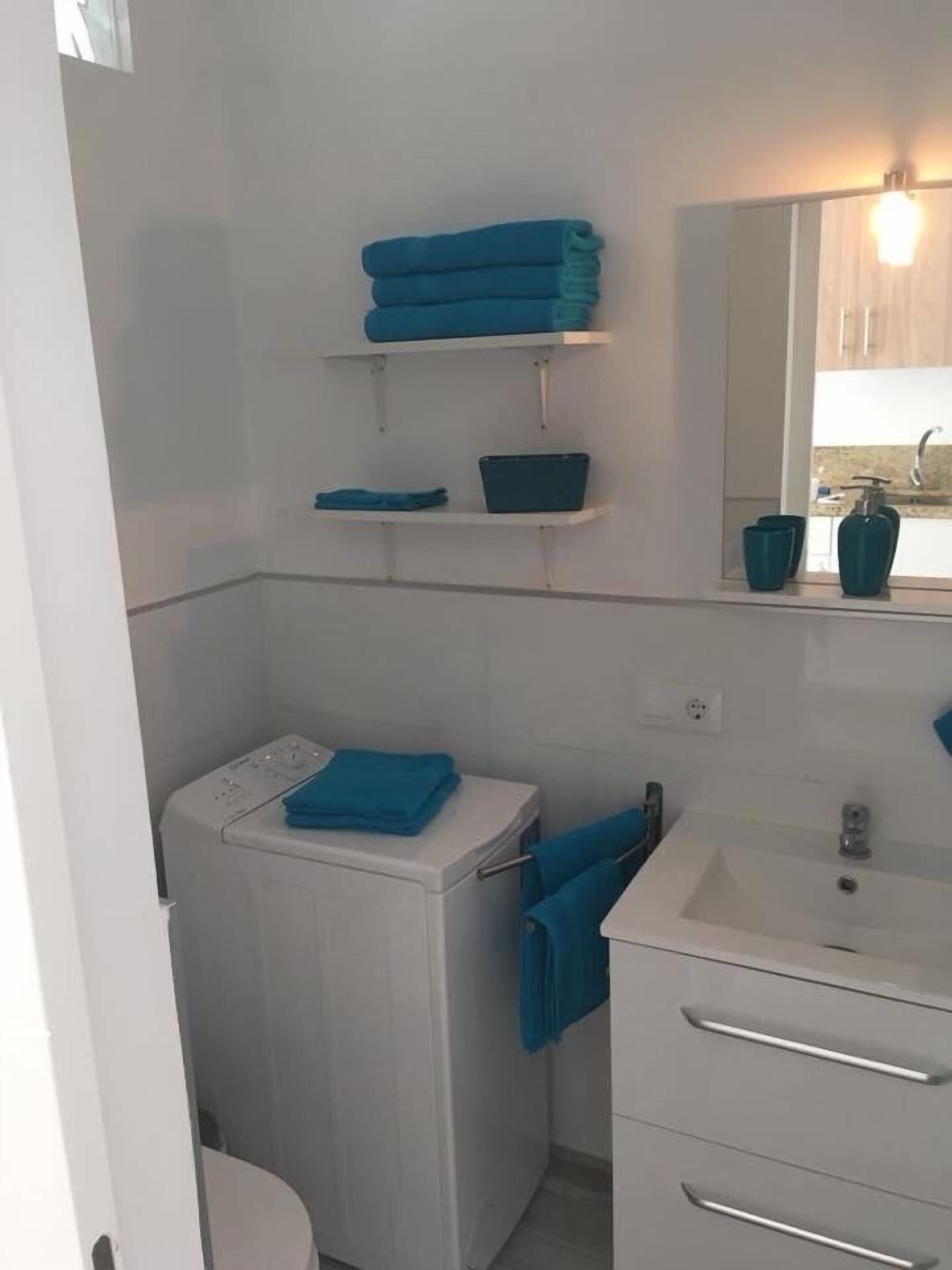 Baño compacto con inodoro blanco, lavabo y espejo sobre él. Dos repisas encima del inodoro sostienen toallas dobladas y un pequeño recipiente.