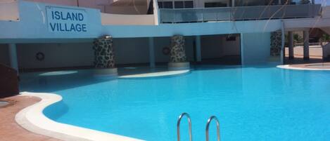 A swimming pool with two metal ladders, surrounded by a tiled deck. A sign reads "ISLAND VILLAGE".