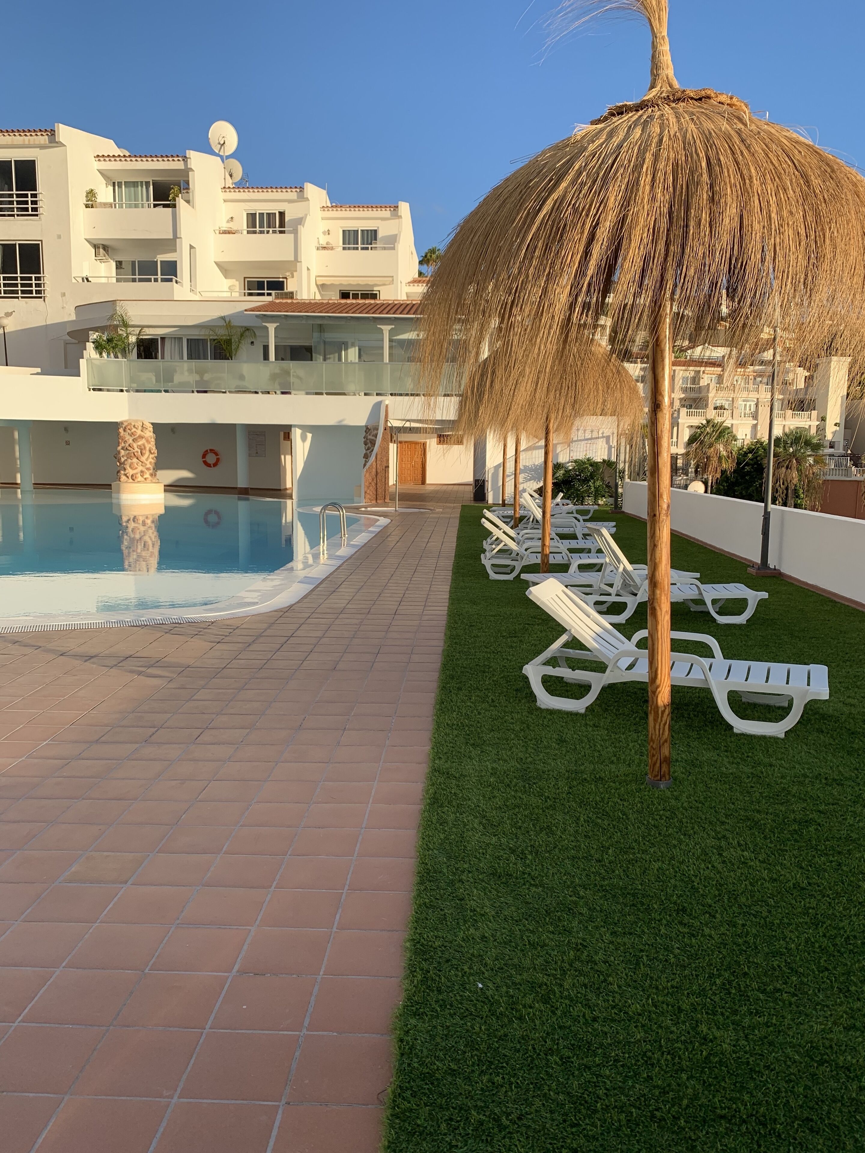 Un espace piscine agrémenté d’un parasol en chaume, de chaises longues blanches et d’un bâtiment doté de balcons et d’antennes paraboliques.