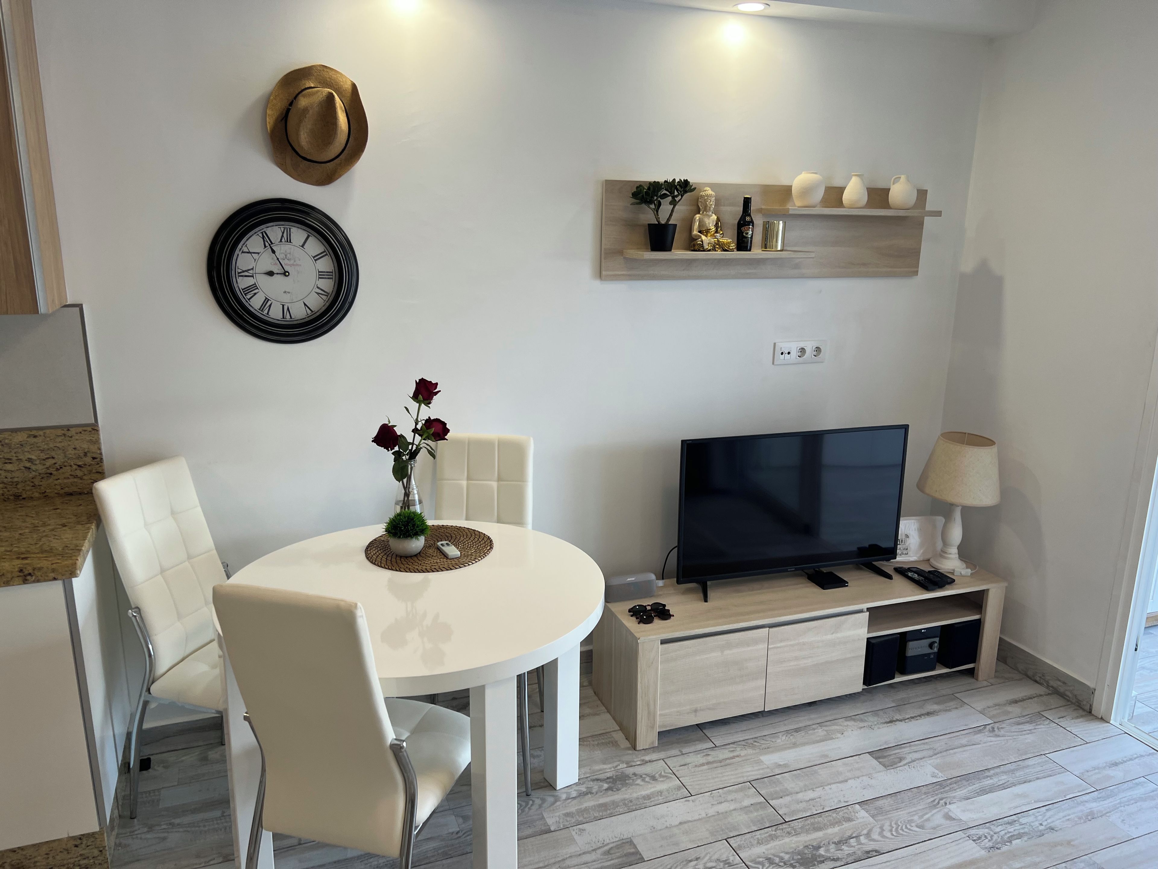 Una sala de estar moderna con una mesa redonda blanca, dos sillas blancas, un reloj de pared, un televisor sobre un mueble de madera y una estantería con objetos decorativos.