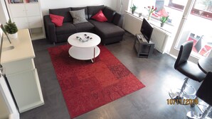 Un salon moderne avec un canapé gris foncé, une table basse blanche et un tapis à motifs rouges. On y trouve une bibliothèque remplie de livres et d’objets décoratifs, un grand tableau représentant une rose rouge accroché au mur, et une fenêtre donnant sur un espace extérieur recouvert d’un tapis rouge.