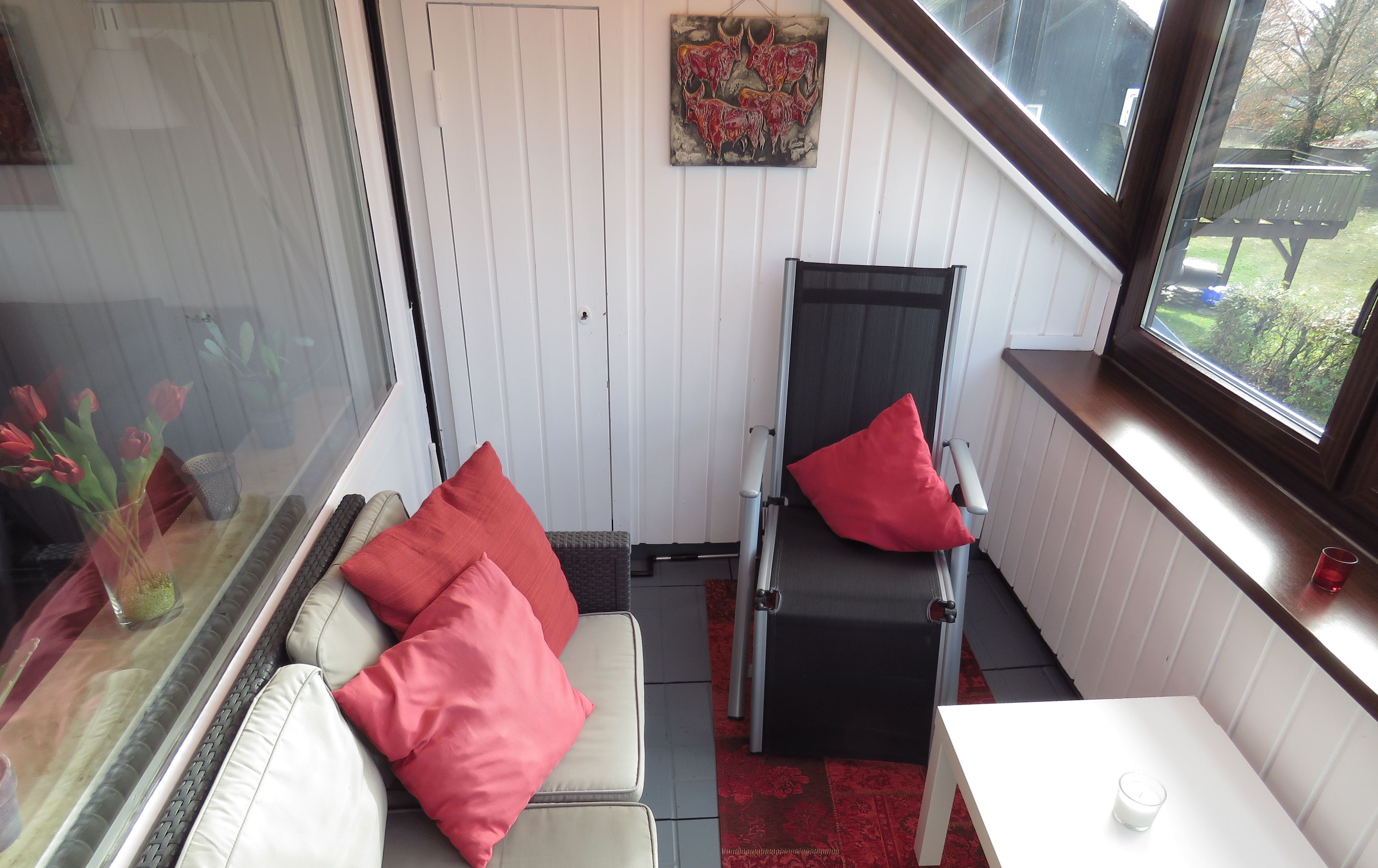 A small room with a white sofa, a black chair with red cushions, a small table with a candle, and a window with a view of the outside.