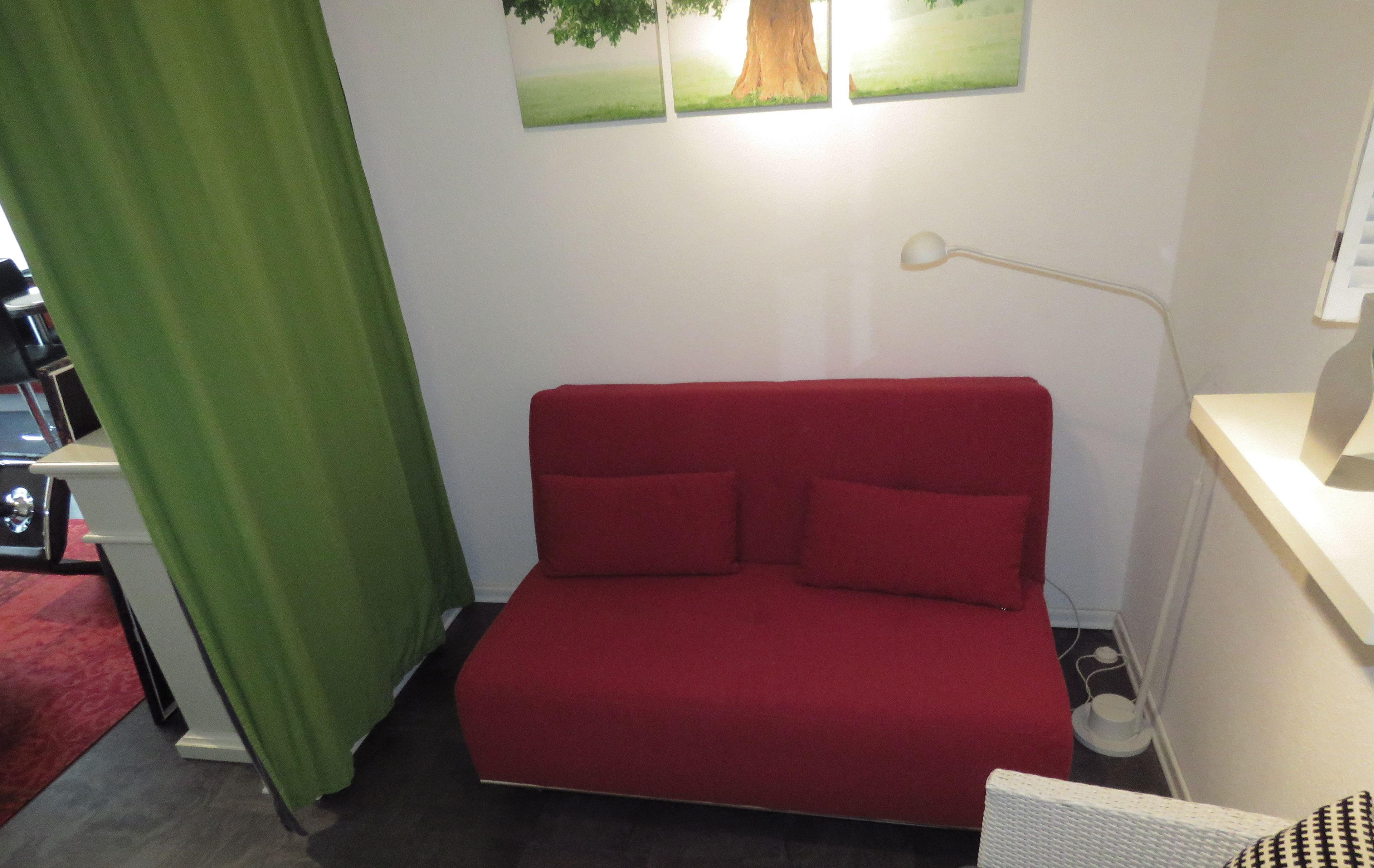 Ein rotes Schlafsofa in einem Raum mit einem grünen Vorhang, einer weißen Lampe und einem Baumgemälde an der Wand.