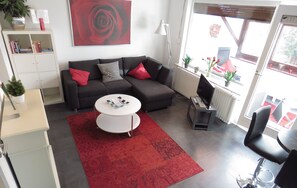 Un salon moderne avec un canapé gris foncé, une table basse blanche et un tapis à motifs rouges. On y trouve une bibliothèque remplie de livres et d’objets décoratifs, un grand tableau représentant une rose rouge accroché au mur, et une fenêtre donnant sur un espace extérieur recouvert d’un tapis rouge.