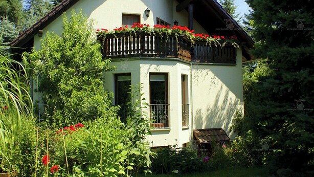 A two-story house with a balcony adorned with red flowers, surrounded by lush greenery. - Quiet holiday home, high quality, lovingly furnished (Dresden)