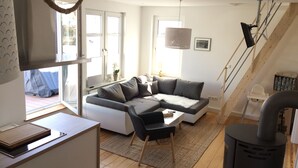 A modern living room with a grey sectional sofa, a small table, and a black chair. There is a large window with a view of the outside, a hanging light fixture, and a staircase leading to an upper level.