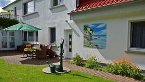 A building with a red-tiled roof, a mural of a coastal scene, a patio with outdoor furniture, and a garden with flowers. - Spacious apartment for up to 5 people centrally on the island of Rügen (Bergen auf Rügen OT Ramitz)