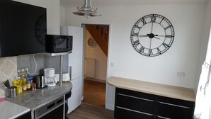 A compact kitchen with a microwave, toaster, and coffee maker on the counter, a large wall clock, and a staircase leading to another room. - 80 m² 100 m from the beach (Hermanville-Sur-Mer)