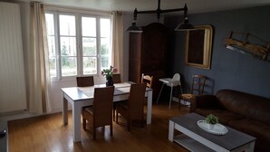 A dining area with a table, chairs, and a vase of flowers. There is a window with curtains, a wall-mounted lamp, and a framed picture. - 80 m² 100 m from the beach (Hermanville-Sur-Mer)