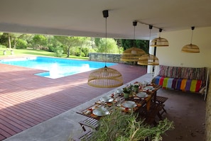 A poolside dining area with a wooden deck, a table set for dining, and hanging wicker lamps.