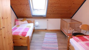 A cozy attic bedroom with a sloped wooden ceiling, a skylight, two wooden beds, a crib, and a radiator.