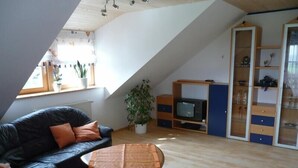 A loft-style living room with a black leather sofa, a wooden coffee table, and a television on a wooden stand. There is a potted plant and a glass cabinet with shelves.