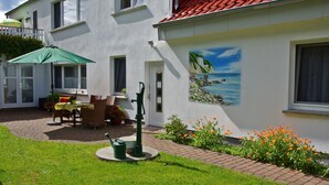 Ein weißes Gebäude mit rotem Ziegeldach, einer Wandmalerei mit Strandmotiv und einer Terrasse mit Gartenmöbeln und einem grünen Sonnenschirm.