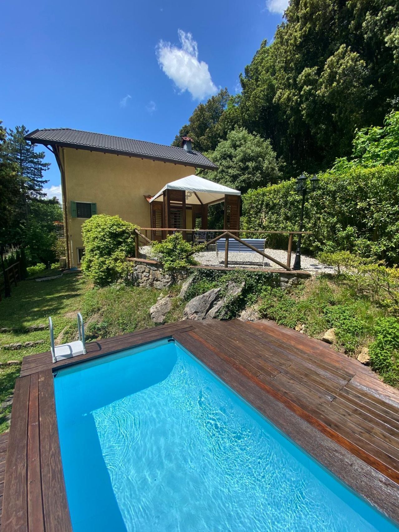 A swimming pool with a wooden deck, a covered outdoor seating area, and a house with a sloped roof.