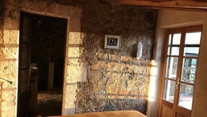 Una habitación rústica con una pared de piedra, una cama con colcha marrón, una puerta de madera y una ventana con contraventanas.