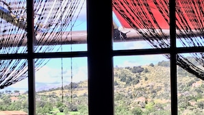 Vista a través de una ventana con una mesa y sillas, que da a un paisaje montañoso con vegetación y algunos edificios.