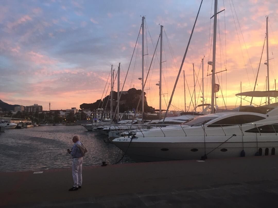 Een jachthaven met meerdere jachten die aanmeren, een persoon op de steiger, en een zonsondergang op de achtergrond.