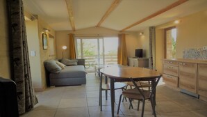 A spacious living area with a sofa, dining table, and chairs, a kitchen with cabinets, and a large window providing natural light. - Stopover 2, in the heart of the Vendée grove (saint florent des bois)
