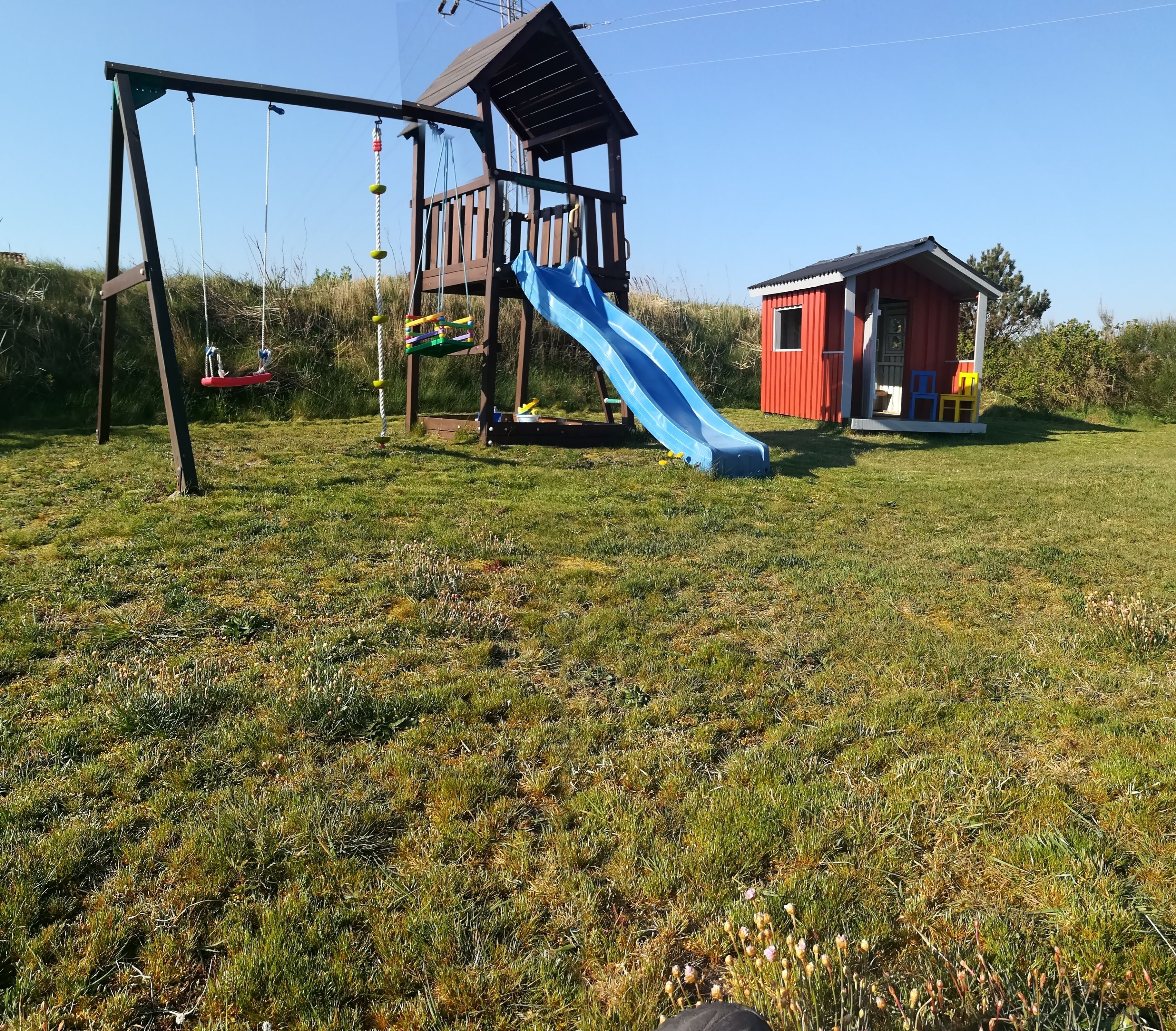 A playground with a swing set and a slide, a small red building, and a grassy field.