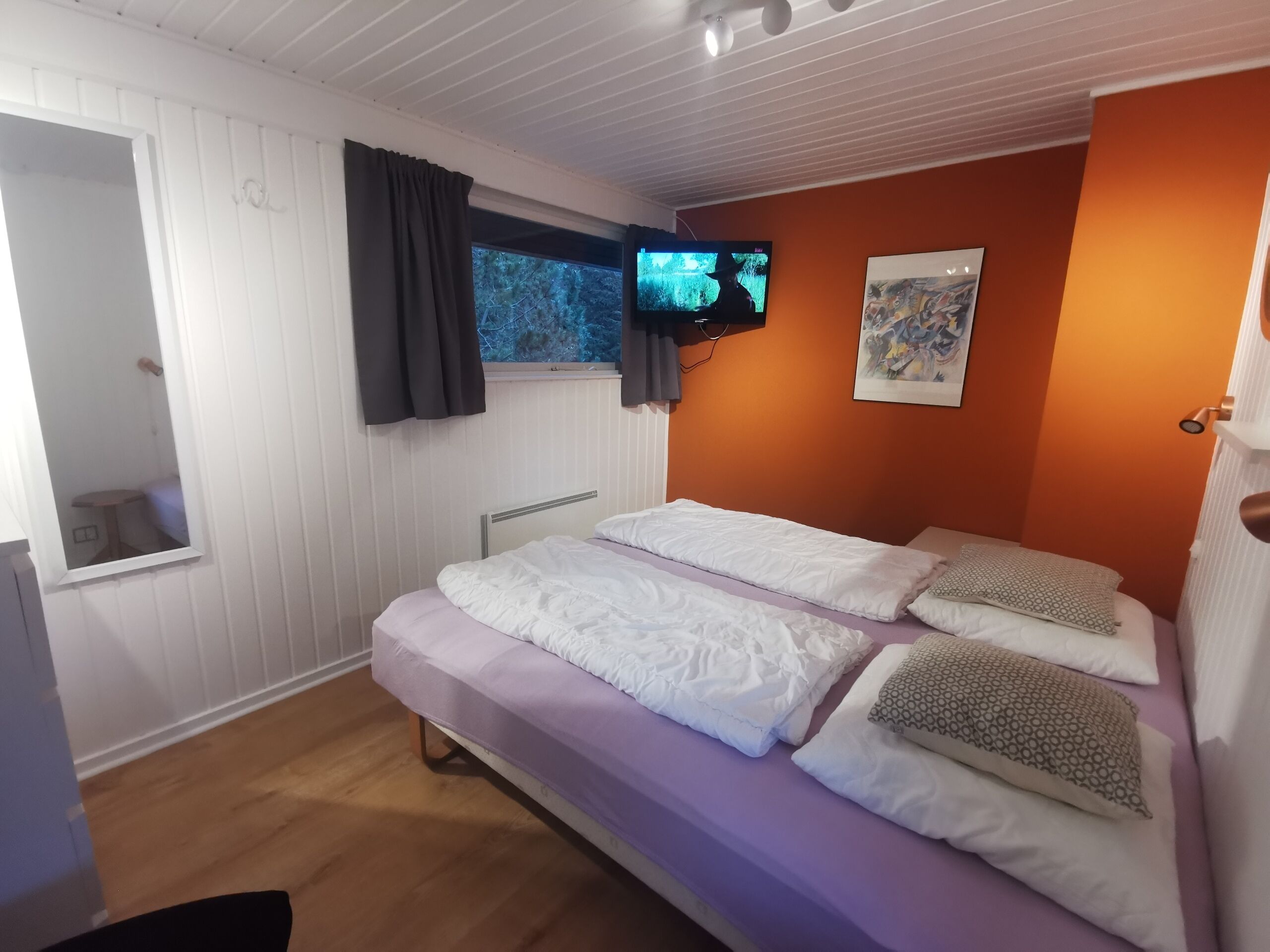 Una habitación con una cama, televisor montado en la pared, ventana con cortinas y un cuadro colgado.