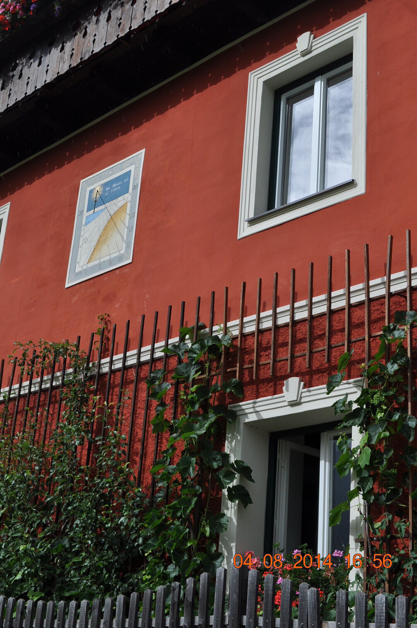 Ein rotes Gebäude mit einem weißen Fenster und einer weißen Tür. Vor dem Gebäude erstreckt sich eine weiße Einfriedung.