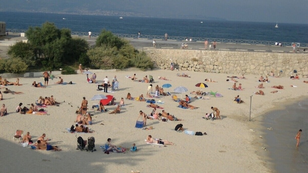 Una playa concurrida, gente tomando el sol y nadando, un rompeolas rocoso y el mar al fondo.