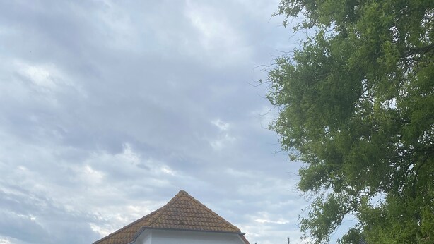 Une maison blanche avec un toit en tuiles, une fenêtre visible et une porte. On y trouve un jardin avec des fleurs roses et un banc en bois.