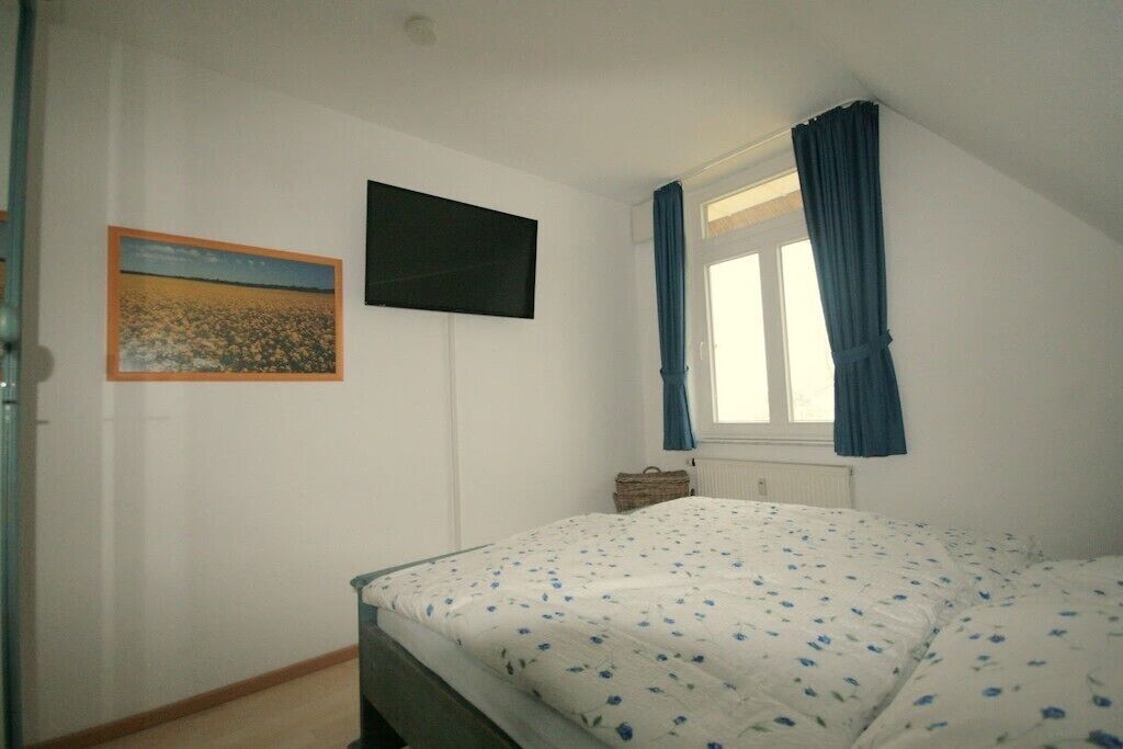 A bedroom with a bed, a window with curtains, a wall-mounted TV, and a framed picture.