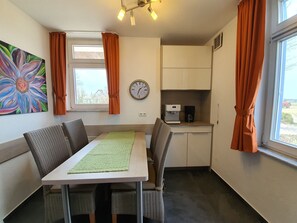 Una habitación pequeña y bien iluminada con una mesa de comedor, sillas, un reloj, una cafetera y una ventana con cortinas.