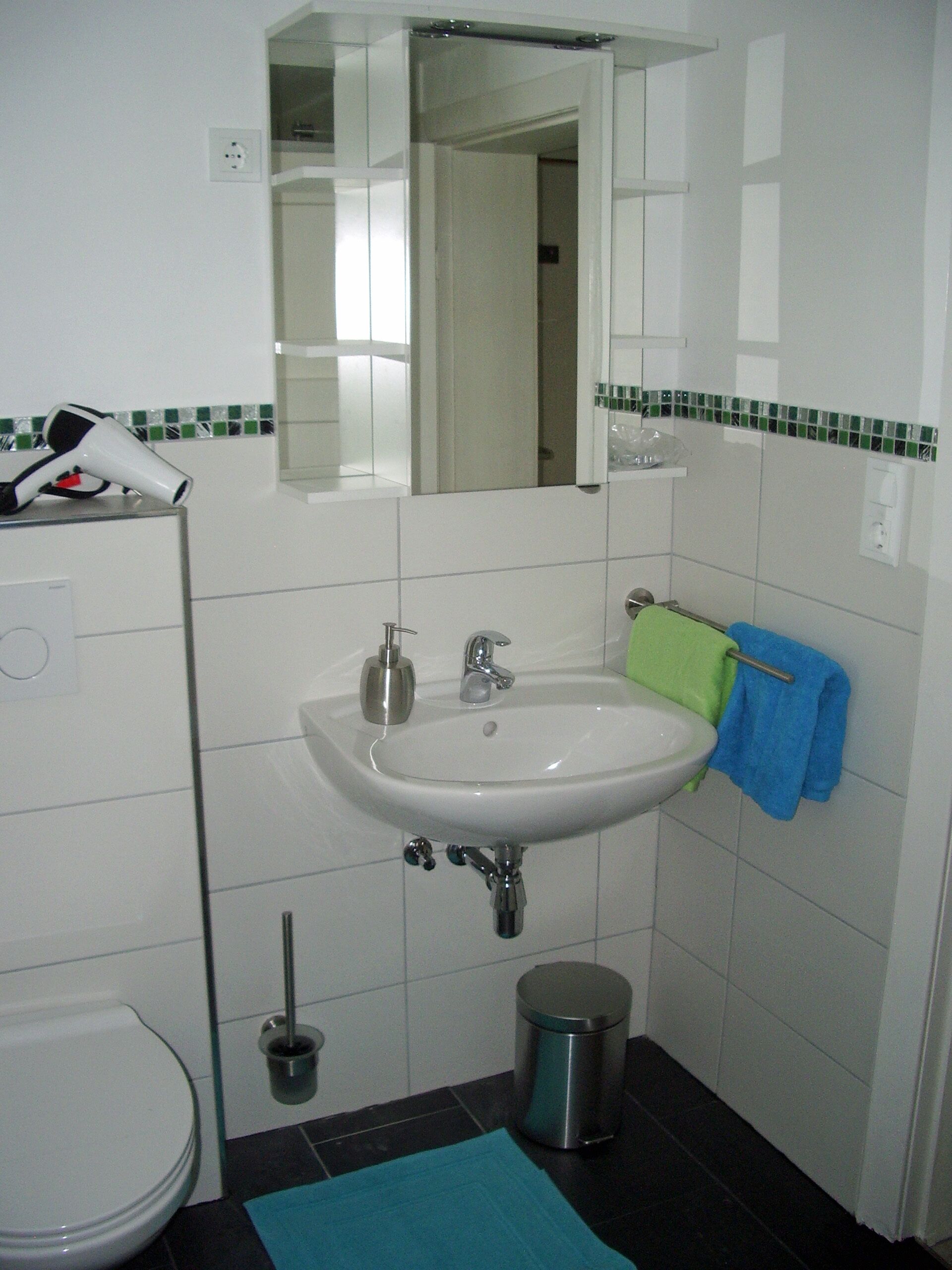 Ein Badezimmer mit weißem Waschbecken, Spiegel und Toilette. Ein grünes Handtuch hängt an der Wand, ein blaues Handtuch liegt auf dem Boden.