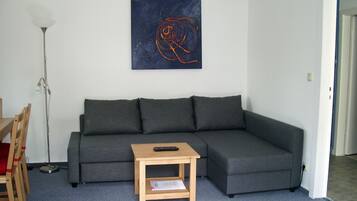 Uma sala de estar com um sofá cinzento, uma pequena mesa lateral de madeira, um abajur e uma obra de arte abstrata na parede.