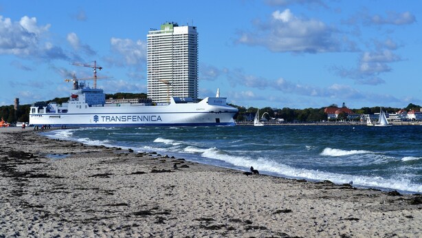 Escena costera con un barco grande llamado 'Transfennica' atracado cerca de una playa, un edificio alto al fondo y un cielo despejado.