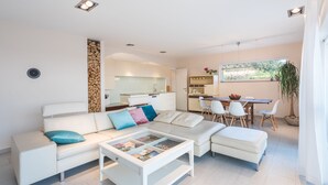 Ein modernes Wohnzimmer mit einem weißen Sofa, einem Couchtisch mit Zeitschriften, einem Essbereich mit Tisch und Stühlen sowie einer Küche im Hintergrund.
