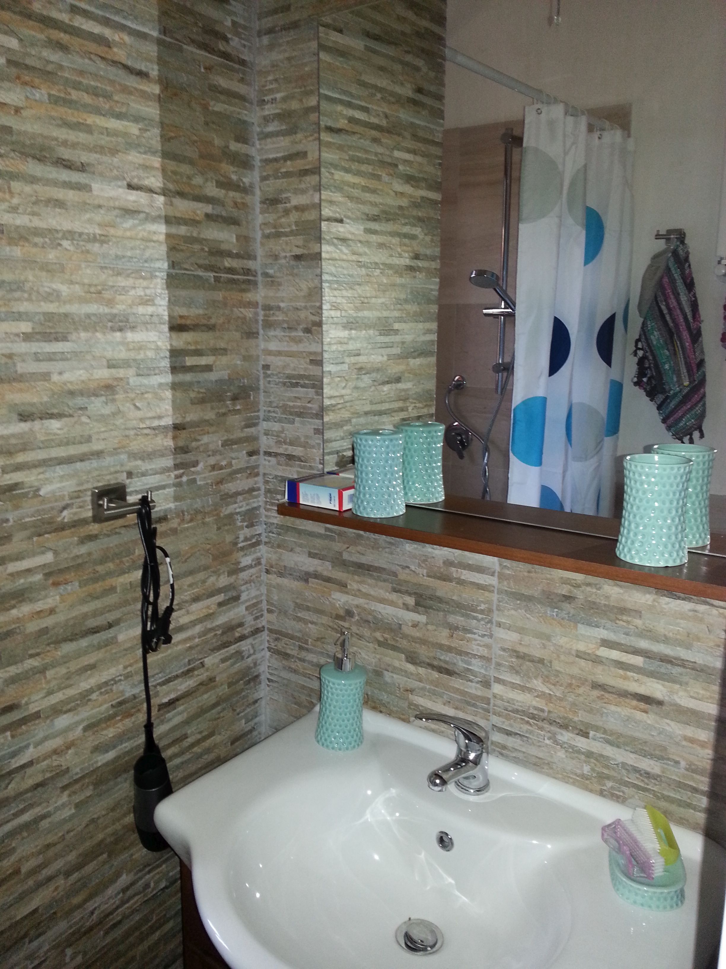 A bathroom with a sink, mirror, and shower. There are toiletries on the sink and a towel hanging on the wall.