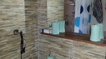 A bathroom with a sink, mirror, and shower. There are toiletries on the sink and a towel hanging on the wall.