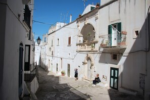 Une rue étroite bordée de bâtiments blancs, un balcon orné de plantes en pot et une personne assise par terre.