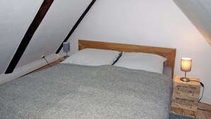Un dormitorio tipo buhardilla pequeño y triangular con estructura de cama de madera, dos almohadas, una lámpara y una caja con la etiqueta 'ESPRIT DE LALANDE' en el suelo.