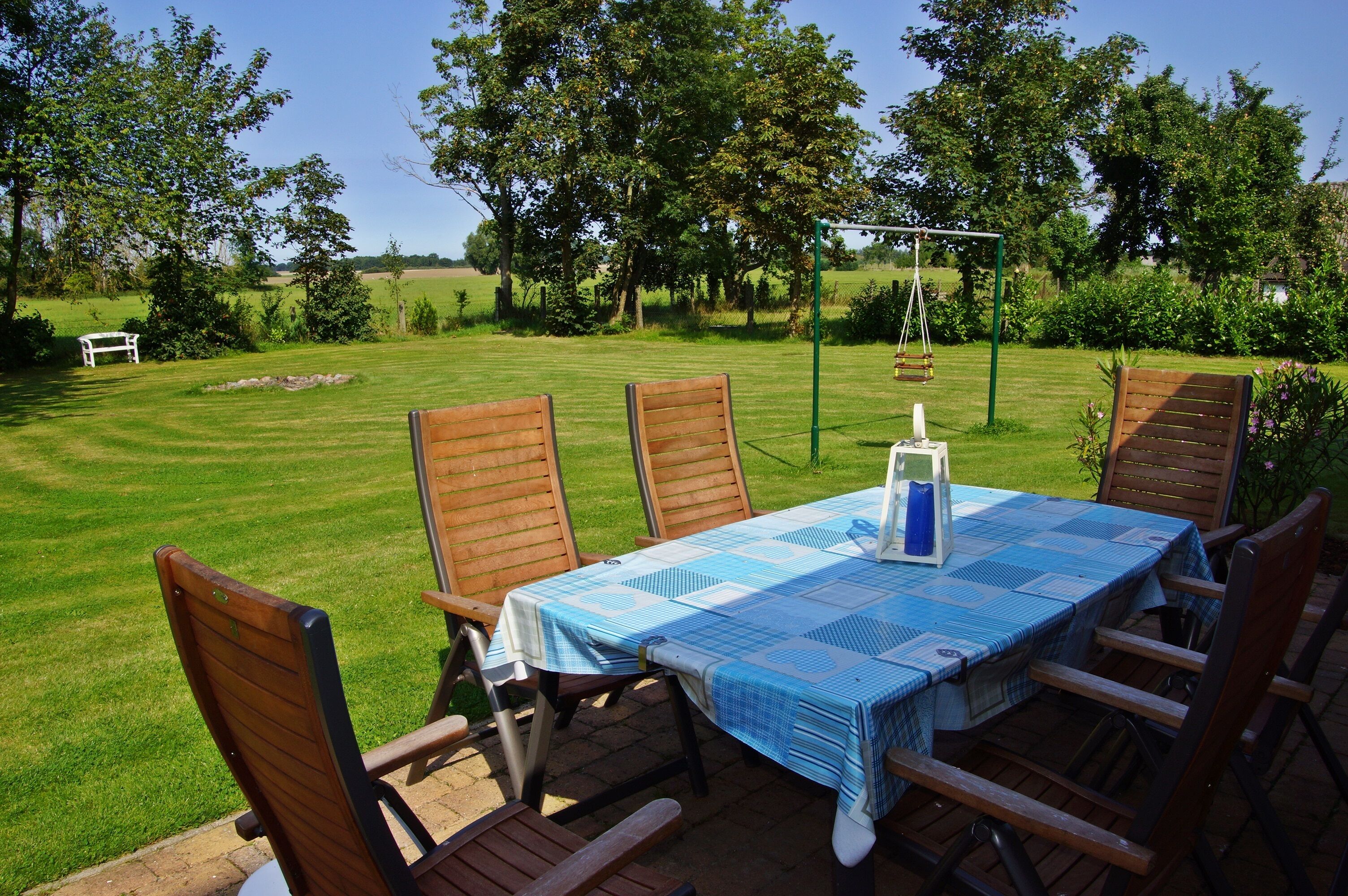 An outdoor dining area with a table set for four, surrounded by wooden chairs, in a lush garden with a swing set and a white bench in the background.