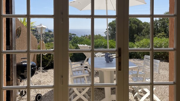 A view through a window with a white frame, revealing a patio area with outdoor furniture, a grill, and a white umbrella. - Typical house of Provence in quiet surroundings, with breathtaking sea-view (Le Lavandou)