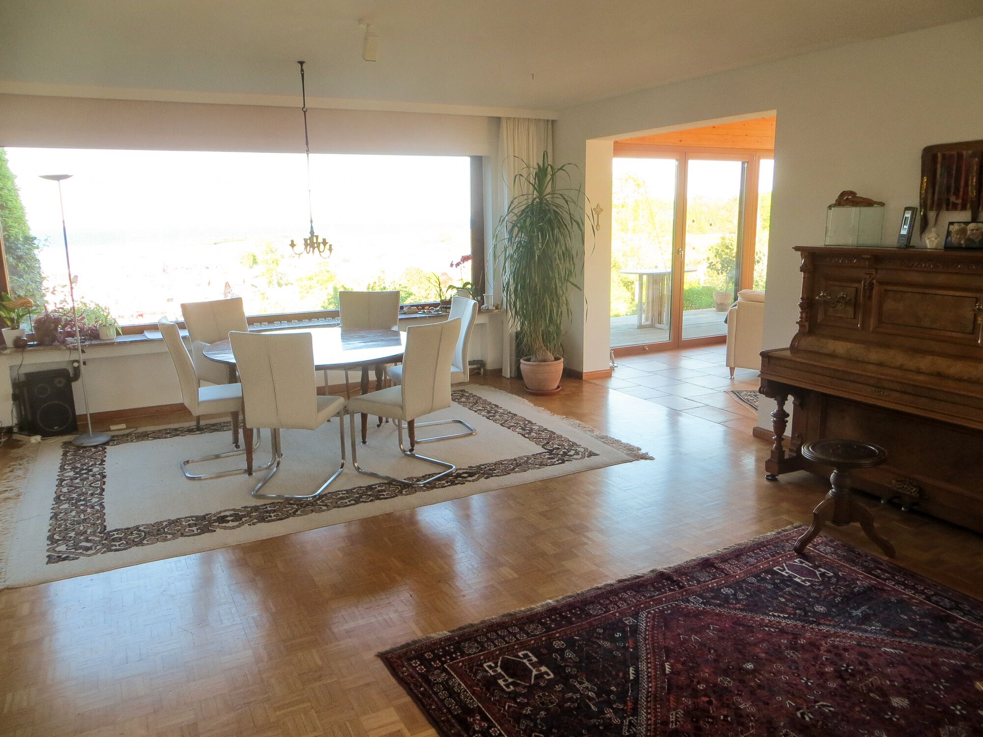 Een ruime woonkamer met een eethoek, een piano en een groot raam dat natuurlijk licht binnenlaat.
