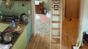 A kitchen with a wooden ceiling, a sink, and a stove. There is a ladder leading to a door.