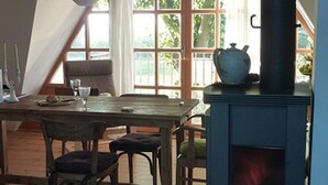 A cozy room with a wooden table, chairs, and a large window with curtains. A fireplace with a visible fire is in the corner.