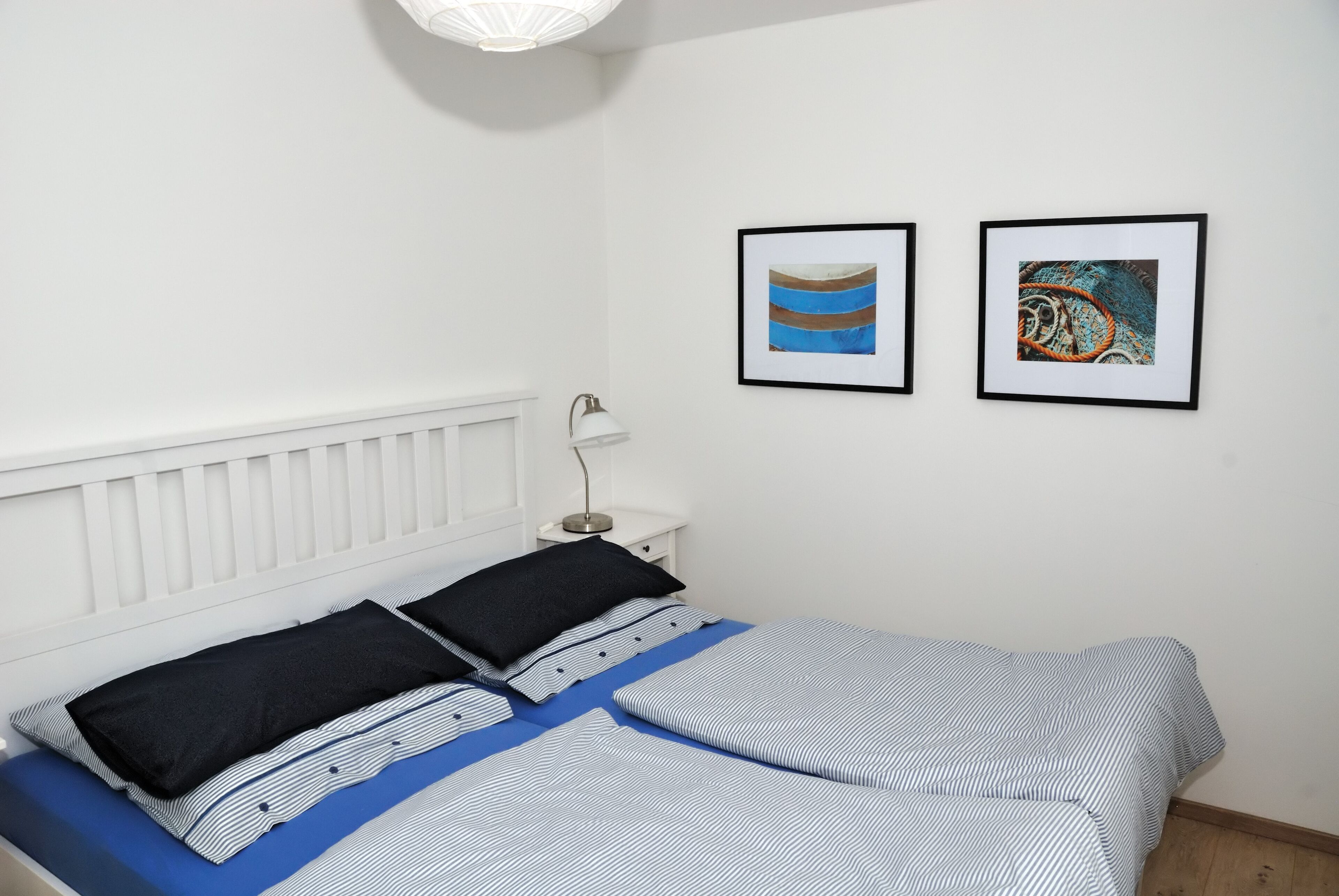 Una cama bien hecha con ropa de cama a rayas, una mesita de noche blanca con una lámpara y dos obras de arte enmarcadas en la pared.