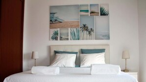 Una cama bien hecha con sábanas blancas, cabecera con almohadones azul turquesa y blancos, y una pared con una obra de arte al estilo playero.