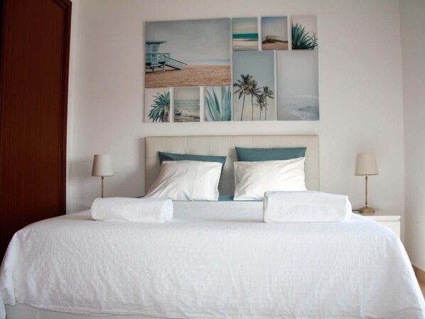 A neatly made bed with white linens, a headboard with teal and white cushions, and a wall with a beach-themed artwork.