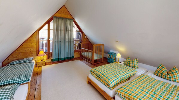 A cozy attic bedroom with a sloped ceiling, two beds with checkered bedding, a crib, a wooden dresser, and a window with curtains.