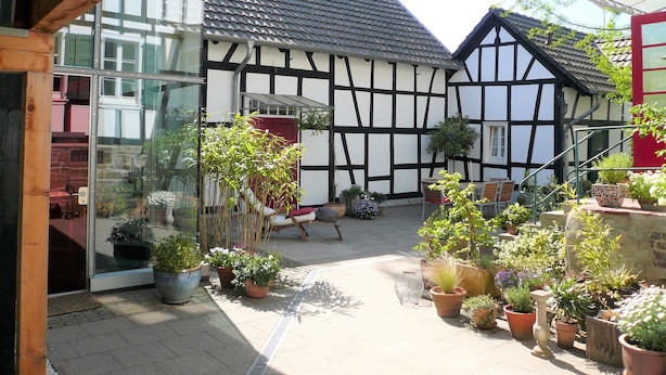 A courtyard with a traditional half-timbered house, potted plants, and a glass door entrance. - Idyllic, completely renovated half-timbered house in a listed courtyard (Neunkirchen-Seelscheid)